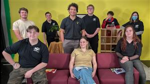 MMT Students sit on or stand on a sofa in front of a yellow wall — the tv studio set